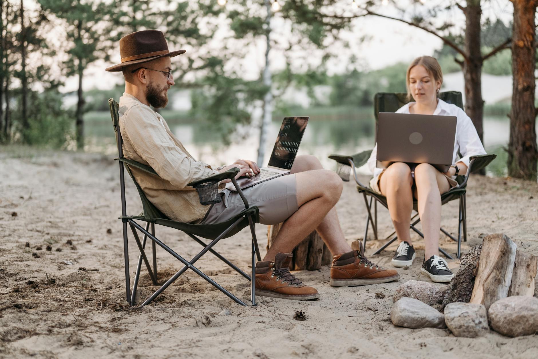 A couple working on laptops at an outdoor campsite table in a natural setting