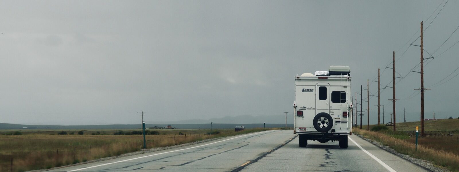 A truck camper on an empty two-lane highway, open plains either side and heavy cloud building ahead.